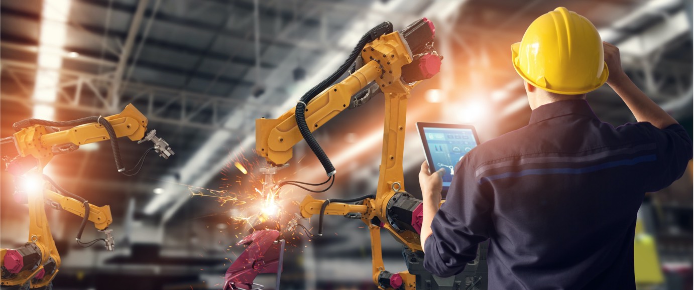 A view from behind of someone wearing a hardhat and holding a digital tablet standing in front of a piece of machinery